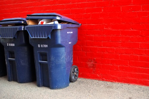 Two large dark blue wheeled rubbish bins are positioned on a paved surface in front of a vibrant red brick wall. The bins have textured plastic surfaces, with lids slightly open revealing some visible rubbish inside. The bins are placed side by side, leaning slightly to the right, with the left bin partially overlapping the right one. The pavement beneath the bins appears to be concrete, and the scene is evenly lit with natural light, giving a clear view of the object details. The backdrop of the red brick wall provides a striking contrast to the dark blue bins, emphasizing their size and form. This setting suggests an outdoor rubbish collection area in a residential or commercial property, consistent with professional waste management services such as those offered by House Clearance Bishops Stortford, which specialise in efficient rubbish removal and clearance services in the local area. The image captures the practical aspect of household or business waste disposal that such companies facilitate.