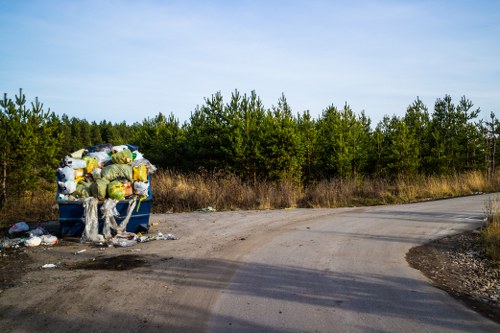 A large blue waste bin situated on a gravel roadside verge near a wooded area, overflowing with a variety of rubbish including plastic bags, cardboard boxes, and miscellaneous litter. The waste appears to be a mix of household waste and general rubbish, with some items spilling onto the surrounding ground. The background features a dense line of green coniferous trees under a clear blue sky, indicating a rural or semi-rural environment. The scene suggests improper disposal of rubbish on a countryside road, which House Clearance Bishops Stortford may address through professional rubbish removal services. The gravel road curves gently to the right, with shadows cast by the trees and vanishing into the distance. The overall setting reflects the casual dumping of waste, contrasting with regulated clearance and recycling services available in the town.
