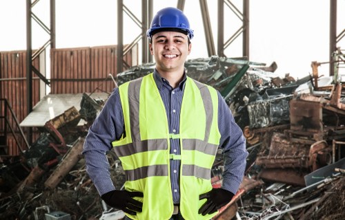 A young male worker in a high-visibility yellow safety vest, blue shirt, black gloves, and a blue hard hat is standing inside a large industrial warehouse or scrapyard. Behind him, there are piles of assorted wrecked vehicle parts, metal scraps, and broken car bodies, many of which are dark grey, black, and rust-colored, with visible textures of damaged metal and worn paint. The background features a structural steel framework and partially open walls, allowing natural light to illuminate the scene. The worker is smiling confidently, with his hands on his hips, indicating readiness for a rubbish removal task. The environment suggests a setting where vehicle disposal or clearance services, such as those offered by House Clearance Bishops Stortford, are carried out, emphasizing the company's local service in the region, possibly near Bishops Stortford or within the wider Hertfordshire area.