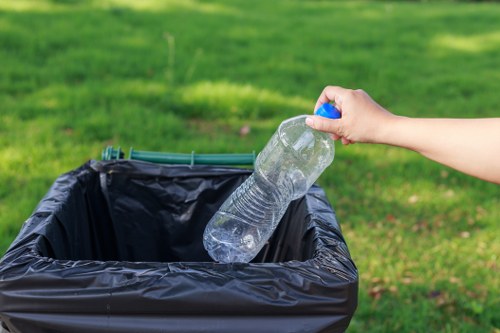 A person's hand is seen holding a clear plastic water bottle over a black plastic rubbish bin lined with a black trash bag. The bottle appears to be partially crushed and is being positioned to discard. The background shows a green grassy area with some patches of sunlight, indicating an outdoor setting. The scene suggests waste disposal activity typical of household rubbish collection, which a company like House Clearance Bishops Stortford may manage. The image emphasizes waste removal and proper disposal of plastic bottles, aligning with services for clearing rubbish from residential or outdoor spaces in the Bishops Stortford area.