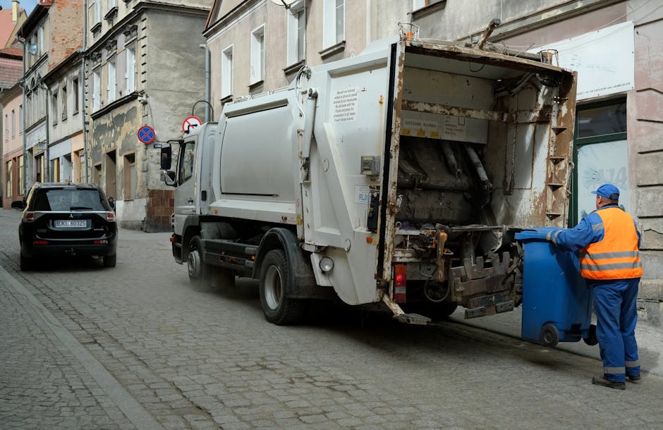 A worker in blue overalls and an orange high-visibility vest is placing a blue wheelie bin into the rear compartment of a white waste collection truck parked on a cobbled urban street. The truck's rea