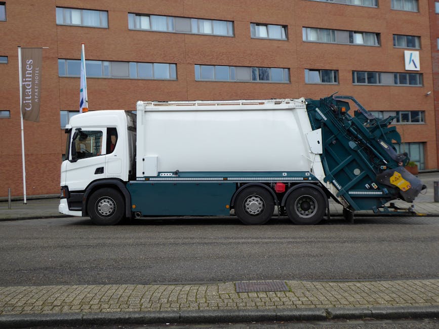 A large industrial rubbish collection truck parked on a city street in front of a modern multi-storey brick building with rectangular windows. The truck features a white cab with a black side mirror a