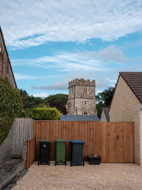 Five black wheeled rubbish bins with yellow lids are lined up against a plain, light-colored wall outdoors, standing on a concrete surface. Each bin has a white oval label with small black text, and t