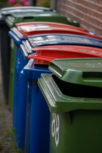 The image shows a residential street with a row of dark grey wheelie bins placed along the sidewalk outside brick houses. A woman dressed in a dark brown coat is seen opening or retrieving items from 